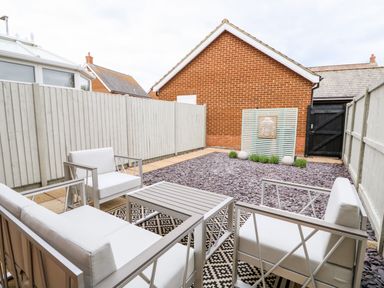 An outdoor patio with white cushioned chairs and a table on a patterned rug in a fenced backyard at 5 Styleman Road in Hunstanton
