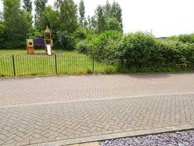 A children's playground with a slide and climbing structure behind a black metal fence and surrounded by green bushes and trees at 5 Styleman Road in Hunstanton
