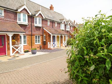 A row of terraced houses with colorful doors and small porches next to a paved street and green bushes at 5 Styleman Road in Hunstanton