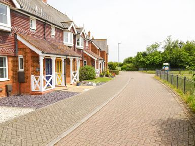 A row of brick houses with colorful doors along a paved street next to a fenced green area at 5 Styleman Road in Hunstanton