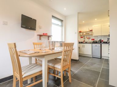 A dining area with a wooden table set with glasses and cutlery next to a kitchen with white cabinets and gray tiled floor at Y Bwthyn Newport in Newport Pembrokeshire