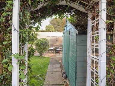 A garden path with a white wooden arch covered with vines leading to a green shed and a patio area with a table and chairs at Y Bwthyn Newport in Newport Pembrokeshire