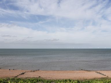 A pebble beach with green vegetation in front and wind turbines in the sea at 4 Country View Park in Graveney near Seasalter