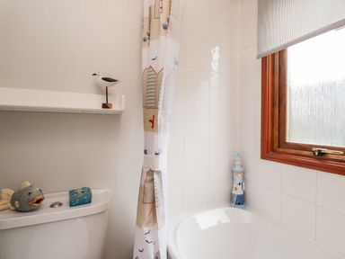A bathroom with a toilet, a white bath with a shower curtain, a window, and decorative items including a wooden bird and a lighthouse at 4 Country View Park in Graveney near Seasalter