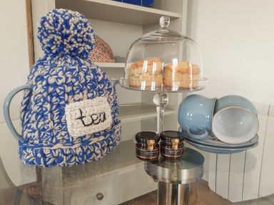 A tea set with a blue and white knitted tea cozy scones under a glass dome jam jars and stacked blue cups and saucers on a glass table at Valley View in Mevagissey