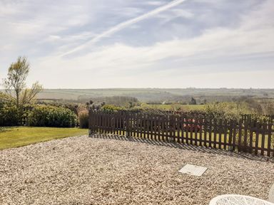 A gravel area with a brown wooden fence and bushes overlooking fields and hills at Valley View in Mevagissey