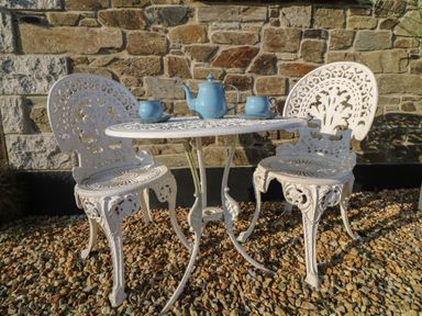 A white metal outdoor table with two matching chairs on gravel in front of a stone wall with a blue teapot and two blue cups and saucers on the table at Valley View in Mevagissey