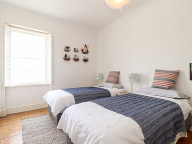 A bedroom with two single beds with striped pillows and blue throws a window with shutters and models of boats on the wall at The Water House in Falmouth
