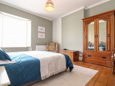 A bedroom with a bed with blue bedding an armchair wooden chest wooden wardrobe and a window with shutters at The Water House in Falmouth
