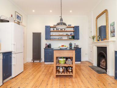 A kitchen with blue cabinets wooden floor a kitchen island and a fireplace with a mirror above it at The Water House in Falmouth