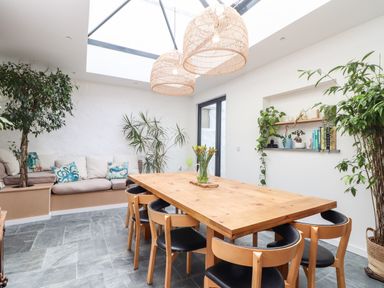 A dining area with a wooden table and six chairs under skylights with plants and a built in seating area at The Water House in Falmouth