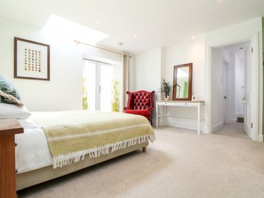 A bedroom with a bed covered with a yellow blanket a red armchair a white desk with a mirror and a plant a door and a window with curtains at The Water House in Falmouth
