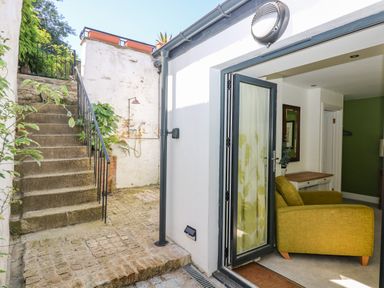 An outdoor stone staircase next to a white wall with plants an open door leading to a room with a yellow armchair and a wooden table at The Water House in Falmouth