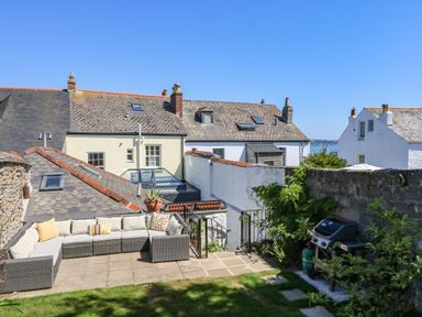 An outdoor patio with a sectional sofa and cushions next to a barbecue grill with houses and a sea view in the background at The Water House in Falmouth
