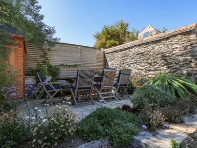 An outdoor patio area with wooden table and chairs surrounded by plants and stone wall at The Water House in Falmouth