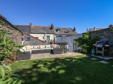 An outdoor patio area with a sectional sofa and barbecue grill in a garden with stone walls and grass at The Water House in Falmouth