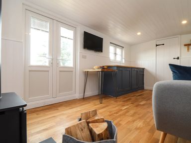 A living room with wood flooring a grey chair a basket of firewood a small table blue kitchen cabinets and French doors at Sebright in St Austell