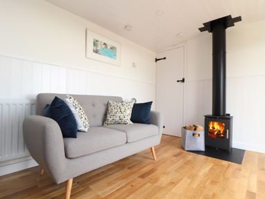 A room with a grey sofa with cushions a wood burning stove and a basket of firewood at Sebright in St Austell