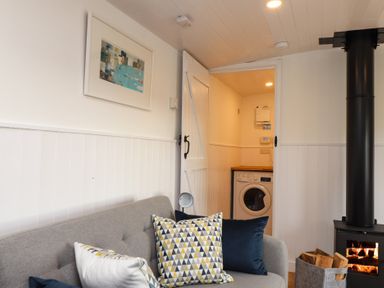 A living room with a gray sofa and pillows near a wood stove and a laundry room visible through an open door at Sebright in St Austell