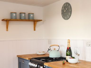 A kitchen corner with a wooden shelf holding three canisters a black stove with a kettle on it a bottle of champagne two glasses and a bowl of strawberries on the countertop at Sebright St Austell