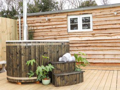 An outdoor wooden hot tub with steps towels and a plant on a wooden deck with a wooden wall in the background at Sebright in St Austell