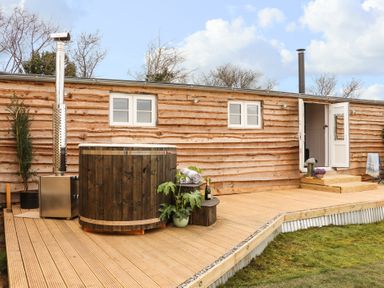 A wooden deck with a hot tub, potted plants, towels, and a bottle next to a wooden building with windows and a door open at Sebright in St Austell