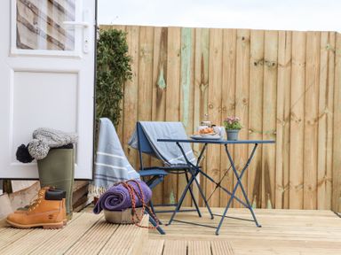 A small outdoor patio with a blue table and two chairs with blankets a pair of boots and green wellingtons next to a door and wooden fencing at Sebright in St Austell