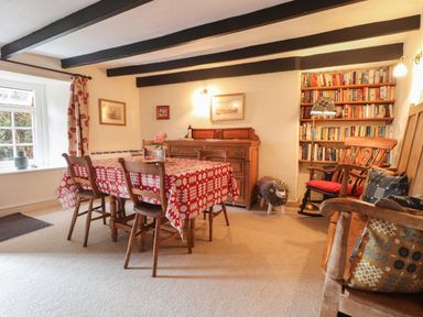 A dining room with a table covered by a red patterned cloth and four wooden chairs a wooden sideboard against the wall a bookshelf filled with books a rocking chair with a red cushion and pillows and a window with floral curtains at Milkwood Cottage in Fishguard Pembrokeshire