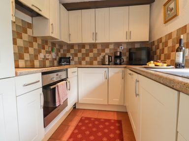 A kitchen with white cabinets a tiled backsplash an oven a kettle a coffee maker a microwave and a red rug at Milkwood Cottage in Fishguard Pembrokeshire