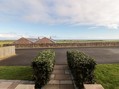 A paved pathway with trimmed hedges on both sides leading to a road with green fields and brick buildings beyond at Low Croft in Embleton