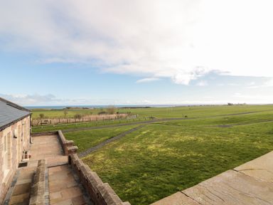 A view of a green field with a stone patio and part of a house with the sea in the background at Low Croft in Embleton