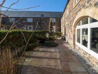 A stone paved patio with shrubs and trees beside a stone house with arched windows at Low Croft in Embleton