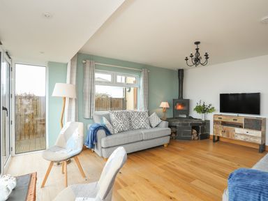 A living room with a grey sofa a wood burning stove two chairs a television and wooden flooring at Red Ridge in Llandanwg near Harlech