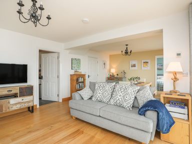 A living room with a grey sofa and patterned cushions a wooden TV stand with a TV an open door a wooden bookshelf a dining area with a chandelier and framed pictures at Red Ridge in Llandanwg near Harlech