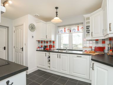 A kitchen with white cabinets black countertops a window with a patterned blind a clock on the wall and various kitchen items on countertops at Red Ridge in Llandanwg near Harlech