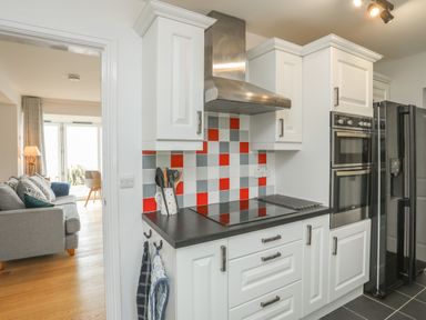 A kitchen with white cabinets black countertop a cooktop and a double oven next to a black refrigerator with a living room visible through an open door at Red Ridge in Llandanwg near Harlech