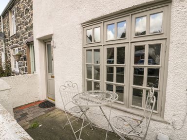An outdoor patio area with a small round metal table and two matching chairs outside a house with a beige door and large multi-pane windows at 3 Alma Terrace in Llanfairfechan
