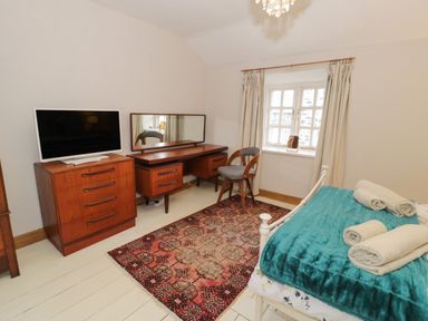 A bedroom with a bed covered with a teal blanket and rolled towels a wooden dresser with a television and a wooden desk with a mirror a chair and a window at 3 Alma Terrace in Llanfairfechan