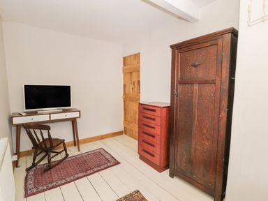A small room with a wooden rocking chair a desk with a television a red chest of drawers a wooden wardrobe and a wooden door at 3 Alma Terrace in Llanfairfechan