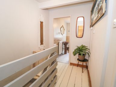 A hallway with a white wooden railing, two mirrors on the walls, a potted plant on a small table, and a view into a bathroom with a brown sink at 3 Alma Terrace in Llanfairfechan