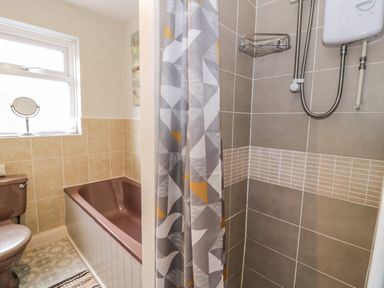 A bathroom with a brown bathtub and toilet a window with a small round mirror a shower area with gray and beige tiles and a patterned shower curtain at 3 Alma Terrace in Llanfairfechan