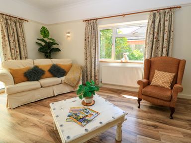 A living room with a beige sofa with cushions a brown armchair a white coffee table with magazines and a plant and floral curtains at Downs View in Norton Ferris