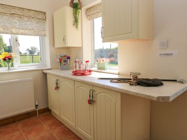 A kitchen countertop with cabinets below and above two windows with shades and various items including glasses flowers and a cane at Downs View in Norton Ferris