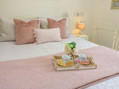 A bed with pink and white pillows a pink quilt two cups with saucers and biscuits on a tray and a bedside table with a lamp at Downs View in Norton Ferris