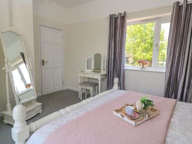 A bedroom with a bed with pink bedding a tray with cups a white dressing table with a stool a full length mirror grey curtains and a window with a view of trees at Downs View in Norton Ferris