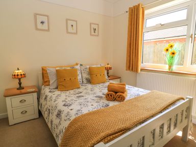 A bedroom with a bed with floral bedding and yellow cushions two bedside tables with lamps three framed pictures on the wall and a window with yellow curtains and flowers in a vase at Downs View in Norton Ferris