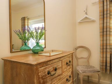 A wooden dresser with a green vase with flowers and a mirror above it next to a chair and window with plaid curtains at Downs View in Norton Ferris