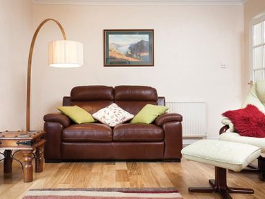 A living room with a brown leather sofa green and patterned cushions a wooden side table a curved floor lamp a framed landscape painting a cream armchair with a red cushion and a matching footstool at Nutwood in Lakeside