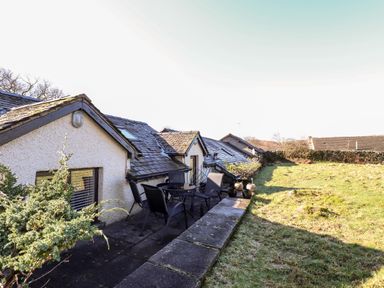An outdoor patio area with a table and four chairs next to a sloped roof house and a grassy yard at Spring Cottage in Ambleside