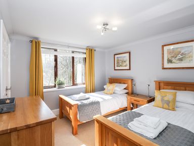 A bedroom with two single wooden beds a wooden dresser a window with yellow curtains and framed paintings at Spring Cottage in Ambleside